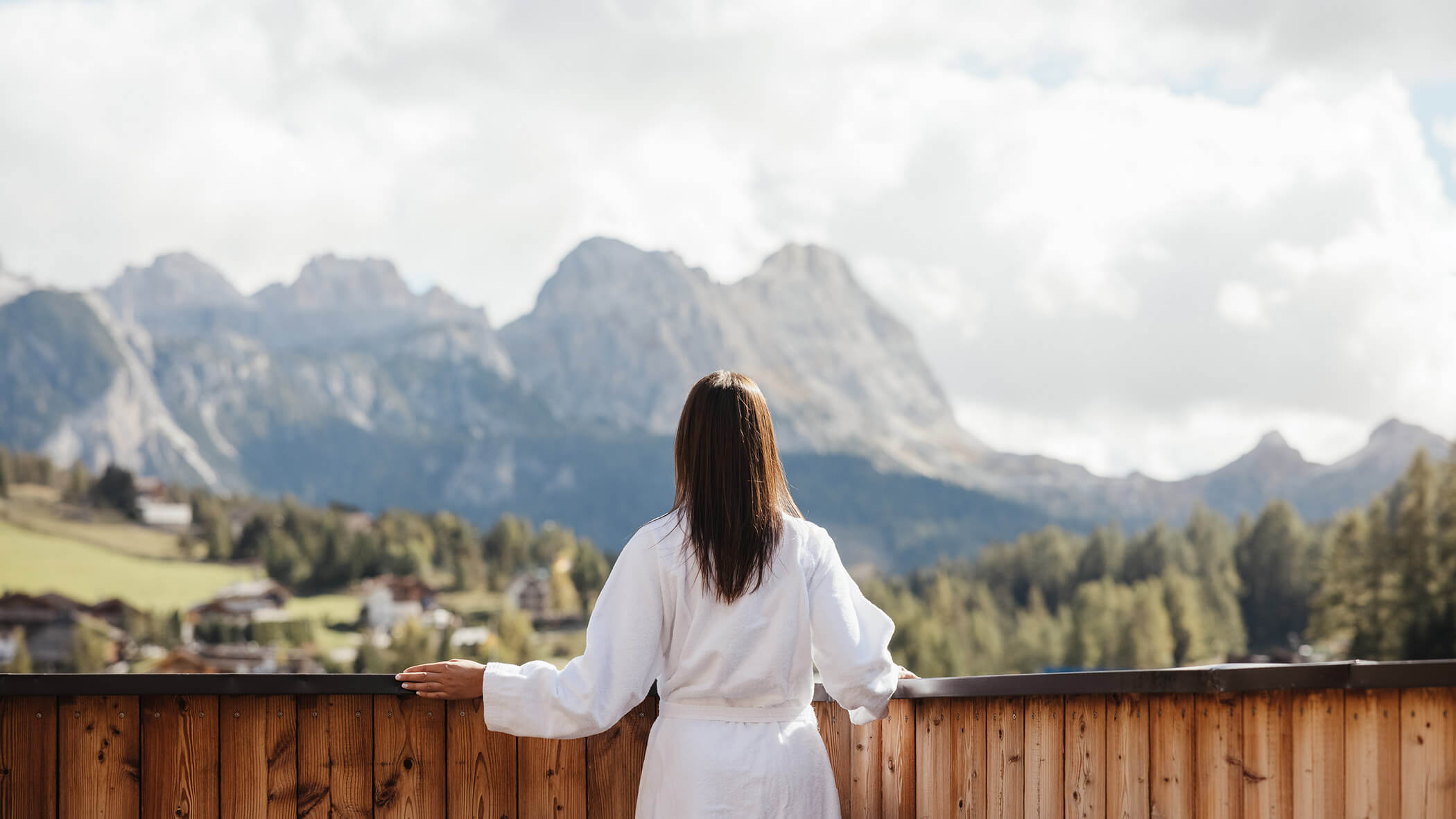 Una donna in vestaglia bianca si trova su un balcone di legno e guarda le montagne lontane e un paesaggio boscoso. - Lagacio Hotel Mountain Residence