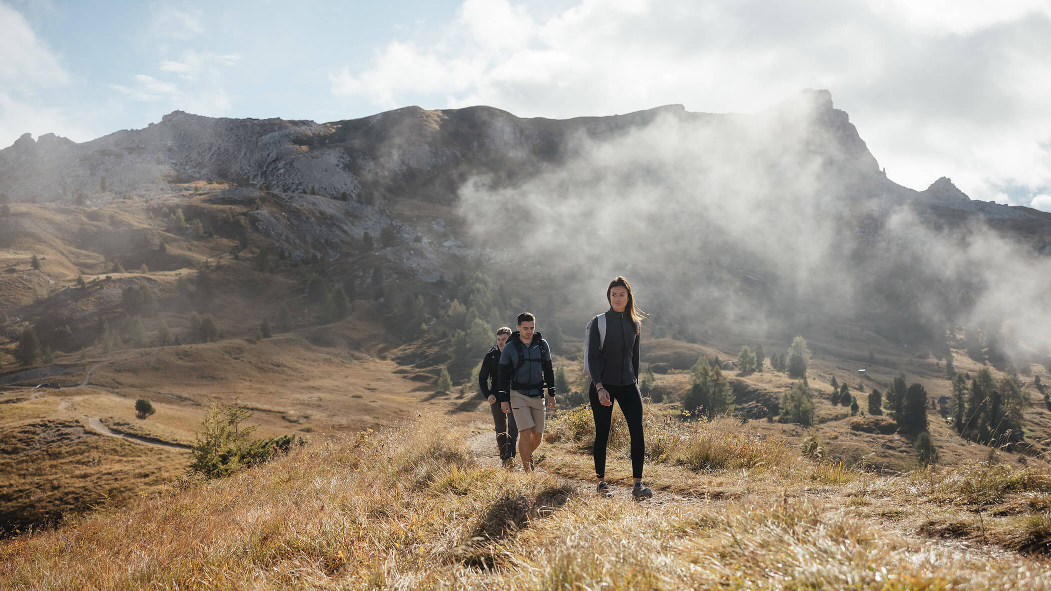 Tre persone percorrono un sentiero erboso di montagna in una giornata nuvolosa con cime rocciose sullo sfondo. - Lagacio Hotel Mountain Residence