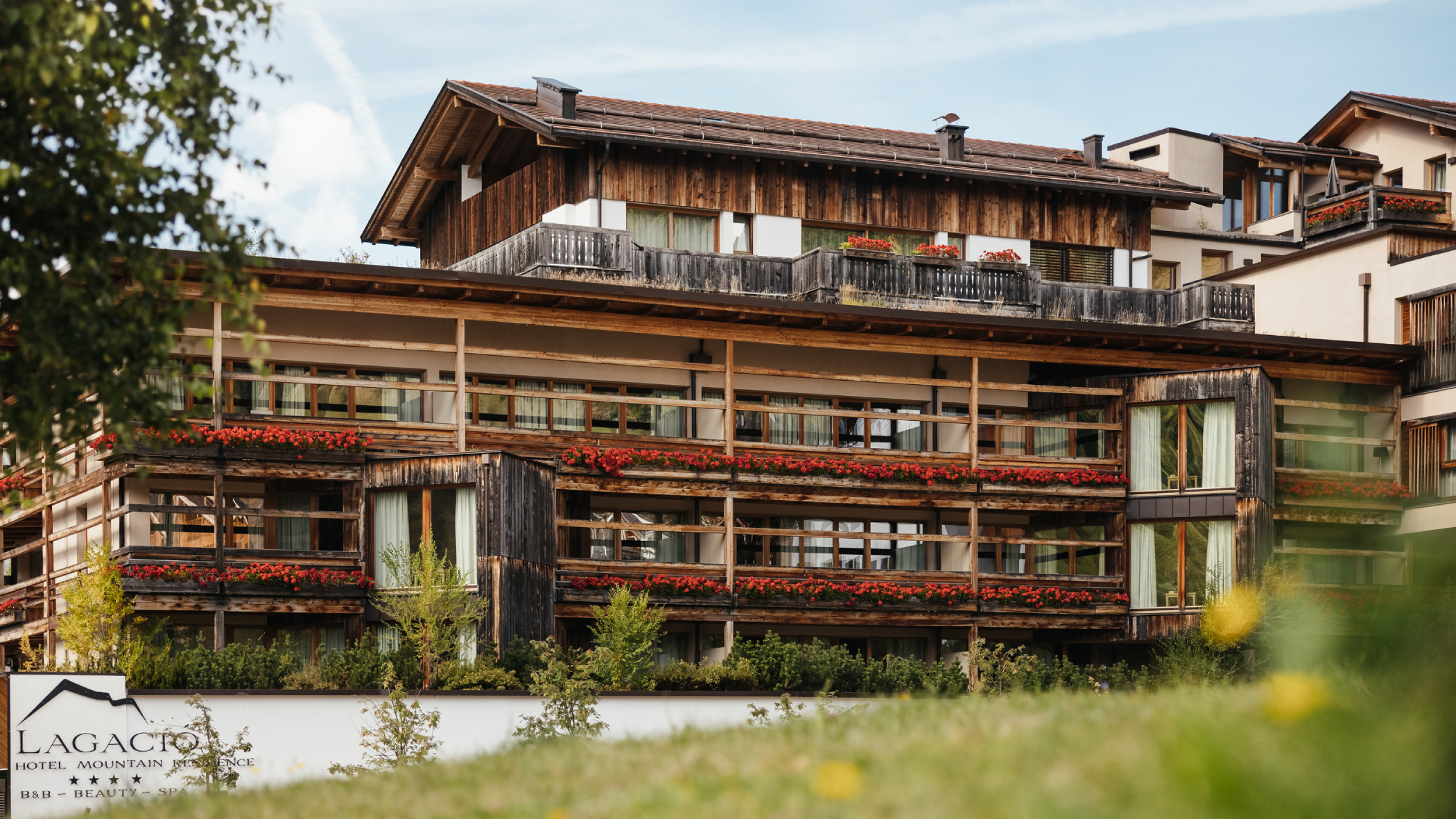 Un hotel alpino in legno con balconi fioriti, immerso nel verde sotto un cielo azzurro. - Lagacio Hotel Mountain Residence